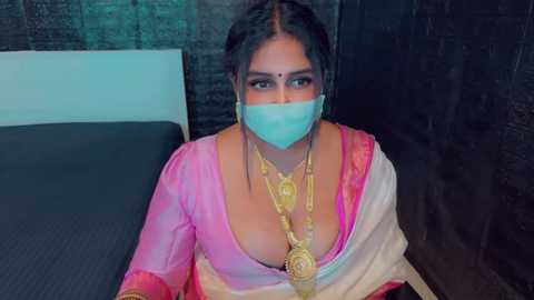 Media: Video of a South Asian woman with dark hair, wearing a pink and white sari, a gold necklace, and a face mask, sitting on a bed against a dark brick wall.