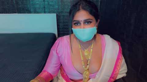 Media: Video of a South Asian woman with medium brown skin, wearing a pink sari with a deep neckline, revealing cleavage. She has a turquoise face mask and gold jewelry, sitting on a bed with a dark headboard.