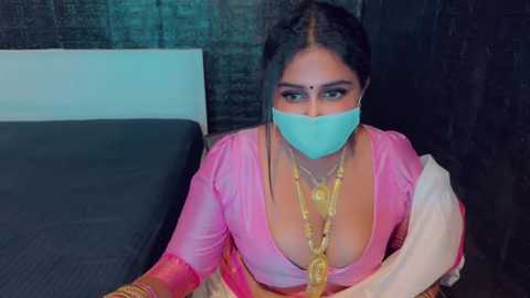 Media: Video of a South Asian woman with long black hair, wearing a pink sari with a deep neckline, gold necklace, and a blue surgical mask, sitting on a bed against a dark wall, with a medical context implied.