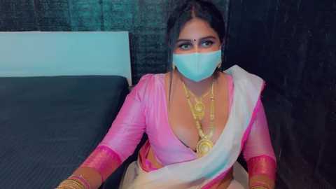 Media: A video of an Indian woman in traditional pink and white saree, gold jewelry, and face mask, seated on a bed with dark walls in the background.