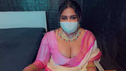 Media: Video of a South Asian woman in a pink saree, adorned with gold jewelry, wearing a face mask, sitting on a dark bed, against a textured black wall.