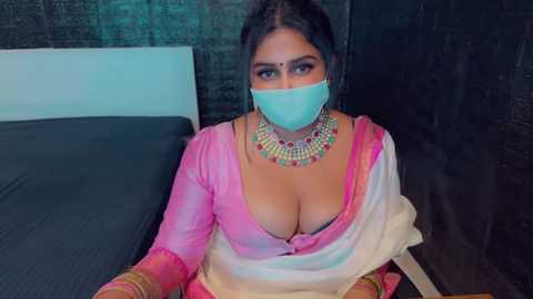 Media: Video of a woman in a pink and white sari with a low neckline, showcasing ample cleavage. She wears a face mask and heavy gold jewelry. Background includes a dark, textured wall and a bed with a black sheet.