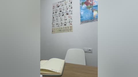 Media: A video of a small, minimalist classroom with a white wall featuring a colorful alphabet poster and a wooden table with a closed book and a white chair.