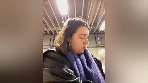 Media: Video of a young Caucasian woman with light skin and shoulder-length brown hair, wearing a black leather jacket and a purple knit scarf, inside a dimly lit, industrial building with exposed wooden beams and fluorescent lights.