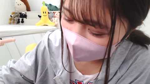 Media: Video of a young girl with long black hair, wearing a light pink face mask and a gray hoodie, sitting in a child-friendly room with stuffed toys and a white desk.