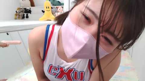 Media: Video of an Asian woman in a white tank top with \"CXX\" written in red, wearing a pink surgical mask, kneeling on a colorful, tiled floor, with stuffed toys and a yellow bear in the background.