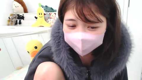 Media: A video of an Asian woman with short brown hair wearing a light pink face mask and a dark gray fur-trimmed jacket, standing in a bright, child-friendly room with white walls, stuffed animals, and a yellow duck toy.