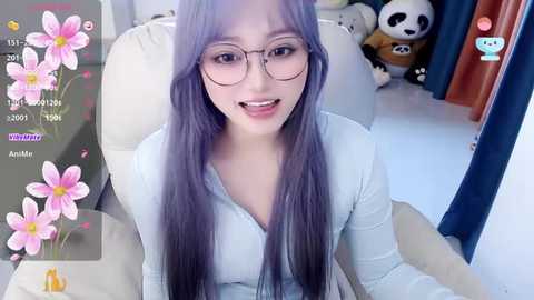 Media: Video of a young Asian woman with long, straight, purple hair, wearing glasses, a light blue V-neck sweater, and a bright smile. Background shows a plush panda and a pink flower filter.