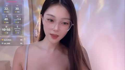 Media: Video of an East Asian woman with long brown hair, fair skin, wearing a light-colored bra, and listening to earphones, indoors with a blurred background.