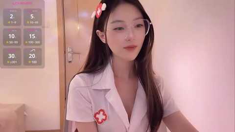 Media: Video of a young Asian woman with long black hair, wearing a white nurse outfit with a red cross emblem, standing indoors near a calendar.