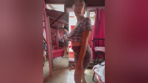 Media: A blurry video of an Asian woman in a tight, patterned crop top and short skirt, standing in a cluttered, red-painted room with a mirror and window.