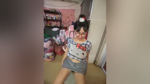 Media: Video of an East Asian woman with glasses, wearing a Hello Kitty crop top and shorts, sitting in a cluttered, pink-themed room with toys, shelves, and a futon.