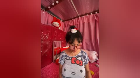 Media: A video of a pregnant woman with glasses, wearing a \"Hello Kitty\" t-shirt, standing in a pink-themed room with Hello Kitty decorations, including a red bear.