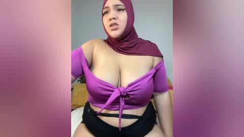 Media: Video of a plus-size woman with a curvy physique, wearing a purple headscarf, off-shoulder top revealing ample cleavage, and black shorts. Background features a plain wall and a bed.