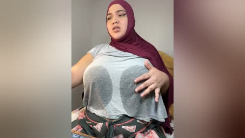 Media: Video of a plus-sized woman with light skin, wearing a gray crop top with a large, visible cleavage, and a maroon hijab. She sits on a yellow chair, wearing patterned pajama pants.