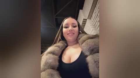 Media: Video of a light-skinned woman with long, straight brown hair, wearing a black low-cut top and a fur coat. She stands in an industrial setting with stacks of white bricks in the background.