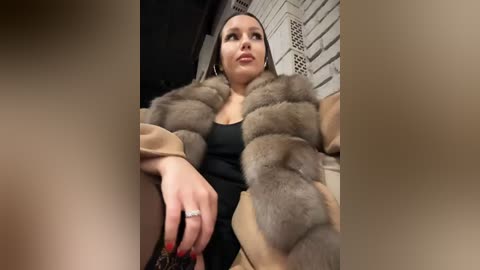 Media: A video of a young woman with light skin and straight, dark hair, wearing a beige fur coat over a black dress, sitting in a brick-walled room with a blurred background.