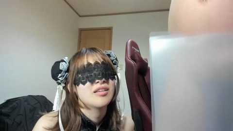 Media: Video of a young Asian woman with long brown hair, wearing a black lace mask and a black and white maid outfit. She is adjusting a red feathered fan in a plain, dimly lit room.