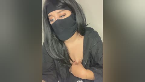 Media: Video of a young woman with long, straight black hair and light brown skin, wearing a black face mask, black hoodie, and a black headband. She is slightly leaning forward, exposing her cleavage.