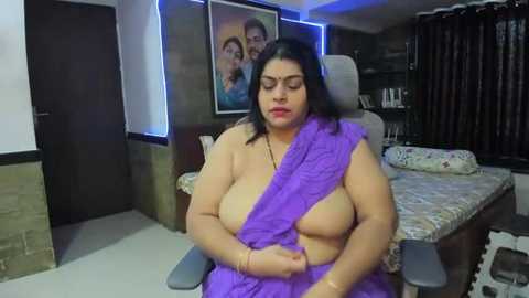 Media: Video of a voluptuous, topless South Asian woman with medium brown skin, wearing a purple sari, sitting on a bed in a dimly lit, modern bedroom.