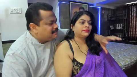 Media: Video of a South Asian couple in a modern bedroom. The man, with a mustache, wears a white shirt, while the woman, with long black hair and glasses, dons a purple saree, gesturing towards a framed photo.