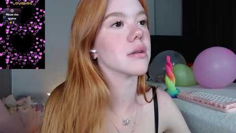 Media: Video of a pale-skinned, red-haired woman with freckles, wearing a black tank top, sitting on a bed with colorful balloons, headphones in. Background includes a black and white patterned wall and a pink keyboard.