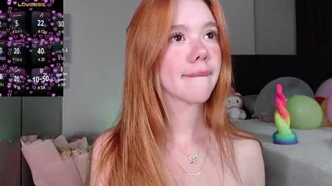 Media: Video of a young, fair-skinned, red-haired woman with a playful expression, wearing a necklace, in a dimly lit room with a pink unicorn toy and a colorful dildo on a bed.