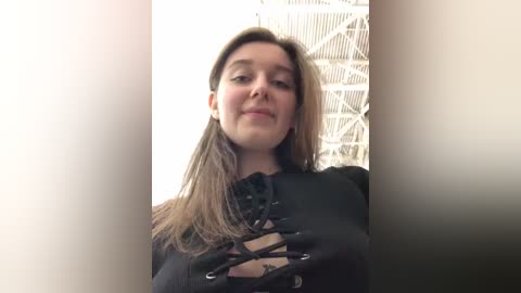 Media: Video of a young woman with light skin, long straight brown hair, wearing a black corset top with lace-up details, standing in front of a white metal structure, possibly a staircase, in a well-lit room.