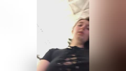 Media: A blurry, low-quality video of a young woman with fair skin and short blonde hair, wearing a black, ripped T-shirt. She's standing in a dimly lit room with white walls and a partially visible white bed.