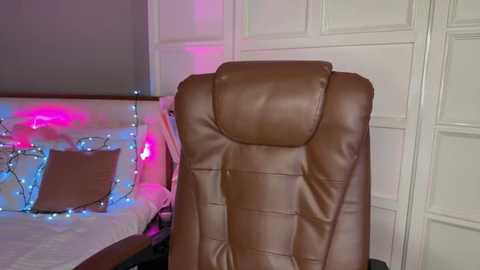 Media: Video of a plush, tan leather office chair with a high backrest, positioned in a modern, minimally decorated bedroom. The background features a bed with white bedding, adorned with a metallic headboard, and a wall-mounted white panel. Soft, multicolored LED lights enhance the ambiance.