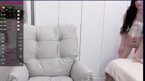 Media: A video of a young woman with long, dark hair, wearing a white lace bra and panties, sitting on a white leather armchair in a minimalist white room.