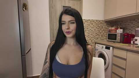 Media: Video of a young woman with long black hair, wearing a blue sports bra and brown jacket, standing in a modern kitchen with beige cabinets, tiled backsplash, and appliances.