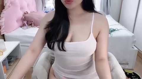 Media: Video of a young woman with long black hair, fair skin, and a slender physique, wearing a sheer white camisole, sitting on a beige chair in a modern bedroom.