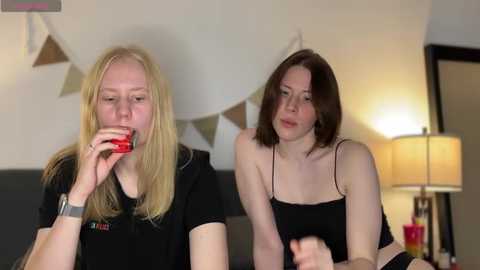 Media: Video of two young women with fair skin and blonde and brown hair, respectively, sitting close together, wearing casual black tops, smiling. Background features a beige lamp, geometric wall art, and a dark couch.