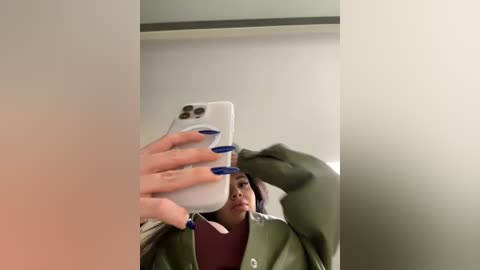 Media: A video of a young woman with dark skin, wearing a green jacket and maroon top, holding a white iPhone with blue nails, taking a selfie in a dimly lit room.