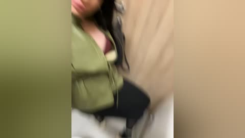 Media: A blurry, low-resolution video of a woman with long, dark hair, wearing a green jacket and white pants, leaning against a beige wall, possibly in a bathroom.