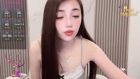 Media: Video of an Asian woman with long black hair, fair skin, and a slender physique, wearing a light gray lace bra, sitting in a modern room with vertical blinds.