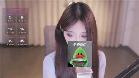 Media: A video shows an Asian woman with long, dark hair in a white shirt, playing a mobile game. A green, cartoonish frog character covers her mouth. The background features beige curtains and a dark floor.