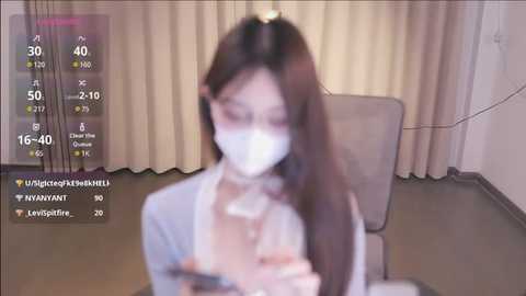 Media: A blurry video of a young Asian woman with long brown hair, wearing a face mask, in a dimly lit room with beige curtains.