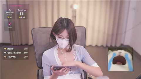 Media: Video of an East Asian woman in a mask, glasses, and light robe, seated at a desk, checking a smartphone. Background shows beige curtains, a lamp, and a small TV screen.