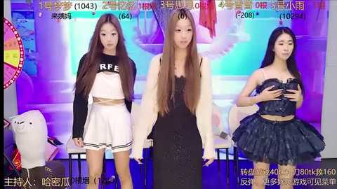 Media: Video of three young Asian women in a studio. One wears a white cheerleader outfit, another a black strapless dress, and the third a black ruffled skirt. Background has colorful posters and a plush toy.