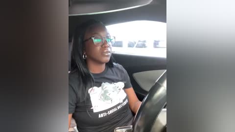 Media: Video of a Black woman with long black hair, wearing glasses, black T-shirt with \"Anti-Racist\" text, and driving a car with a blurred parking lot background.