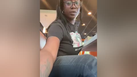 Media: Video of a young Black woman with medium-brown skin, long braids, and glasses, wearing a black graphic t-shirt and jeans, seated in a modern, brightly lit cafe.