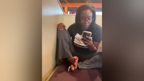 Media: Video of a Black woman with glasses, medium build, sitting on a red mat, wearing a black t-shirt and gray pants, taking a selfie with a smartphone. Background shows a beige wall and a window with orange trim.