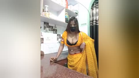 Media: Video of a dark-skinned woman in a yellow sari with a black bra, cleaning a kitchen counter, wearing a face mask. Background includes white cabinets, a sink, and a window.