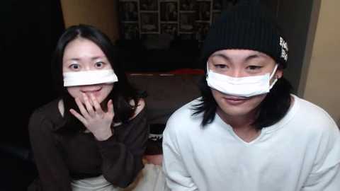 Media: Video of two Asian women wearing white masks, black beanie hats, and dark sweaters, sitting indoors with dim lighting and a cluttered background.