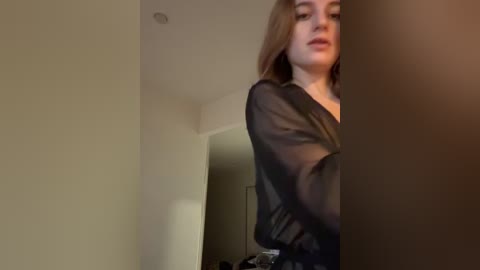 Media: A video of a young woman with light skin and long, wavy blonde hair, wearing a sheer black blouse, standing in a dimly lit room with beige walls and a partially visible door.