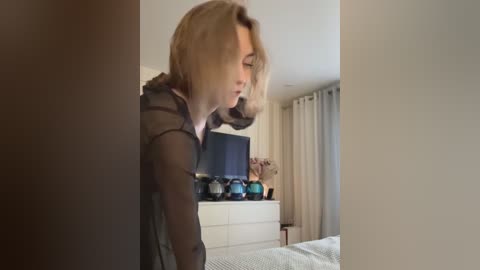 Media: Video of a blonde woman with shoulder-length hair, wearing a sheer black top, leaning over a white dresser in a softly lit bedroom with beige curtains and a flat-screen TV.