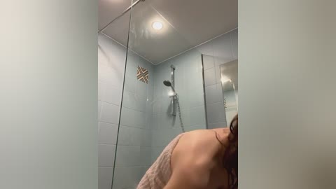 Media: Video of a nude woman with wet, dark hair, standing in a minimalist, light blue-tiled shower. She holds a towel wrapped around her shoulders.