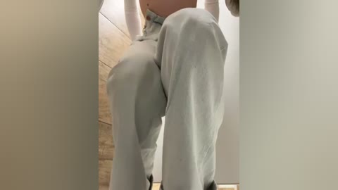 Media: Video of a woman's lower body in light gray pants, viewed from the side, standing in a beige-tiled bathroom with a brick wall in the background.